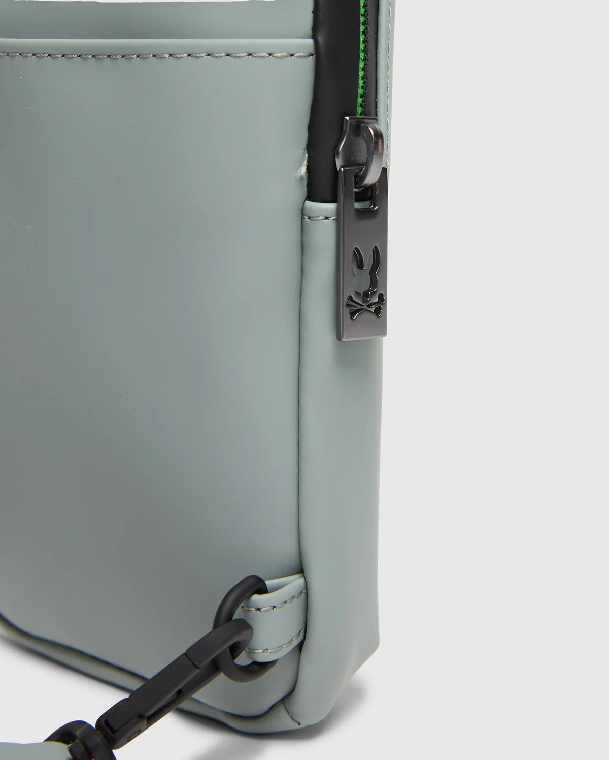 Close-up of the corner of a light gray Psycho Bunny MENS RUBBERIZED CROSSBODY (B6A635F200) crafted from durable polyester, featuring a black zipper with a sleek rabbit logo, visible stitching, and an attached black crossbody strap.