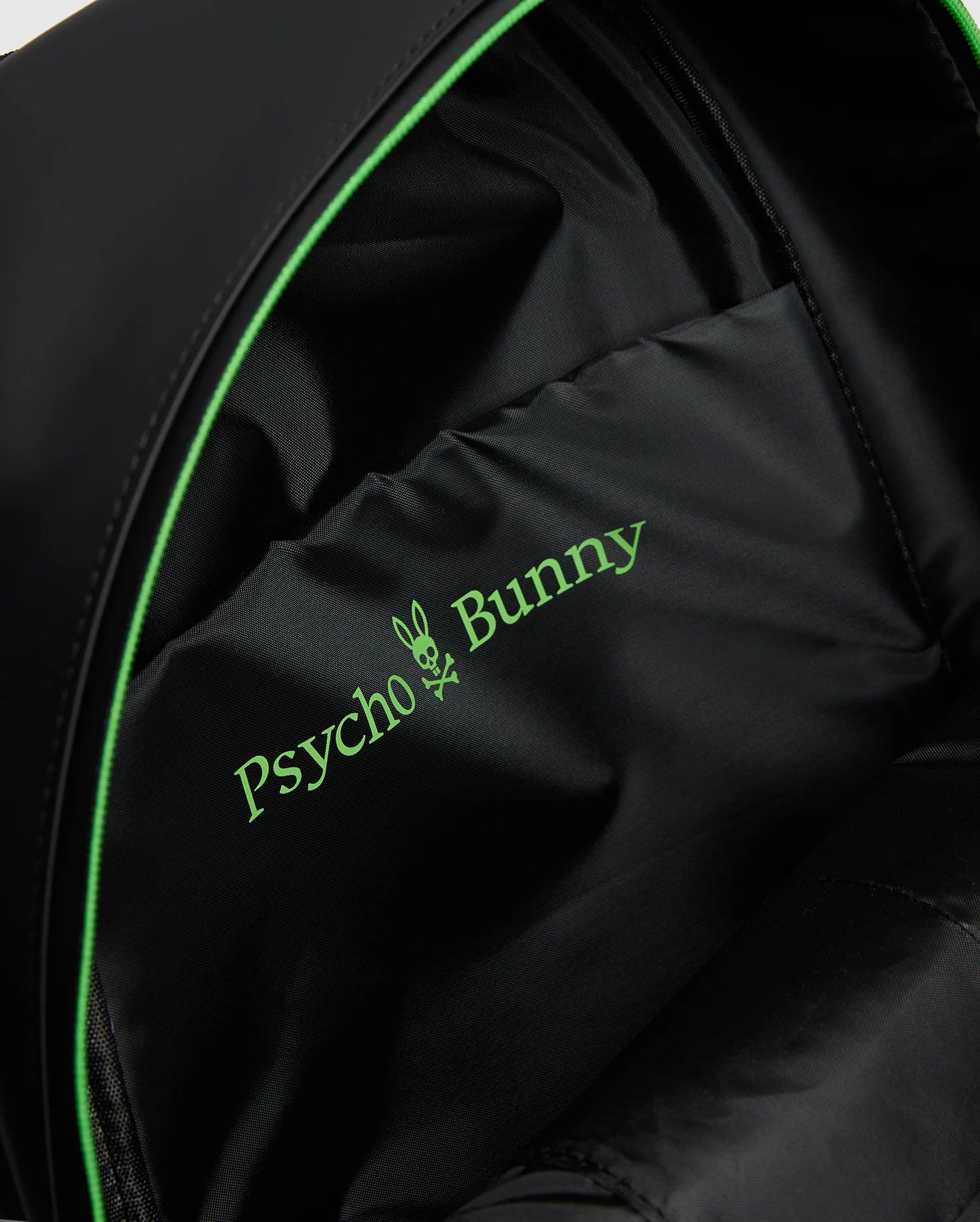 Close-up of the black MENS RUBBERIZED BACKPACK (B6A637F200) interior by Psycho Bunny, featuring a green bunny crossbones logo, showcasing its durable and functional design with robust lining and precise stitching.