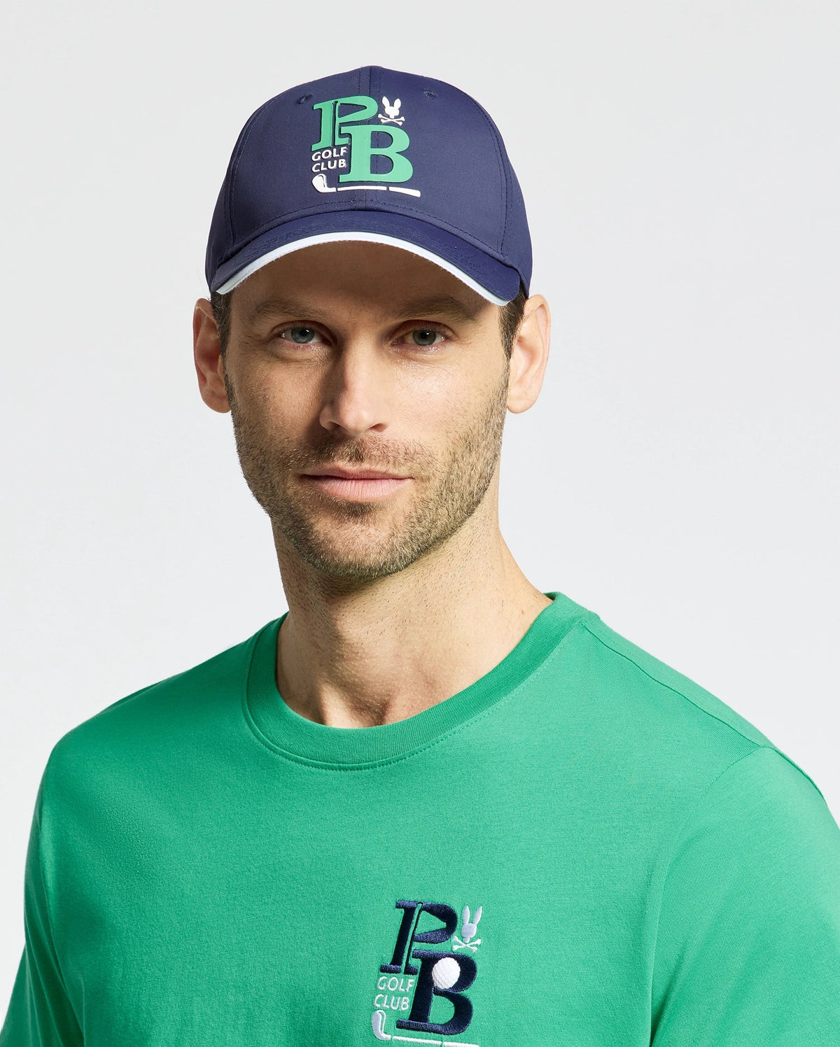 A person wears the Psycho Bunny MENS DENIS SPORT CAP in navy, adorned with a green and white "PB Golf Club" logo, paired with a matching lightweight cotton blend green T-shirt. The background is plain and light-colored.