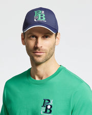 A person wears the Psycho Bunny MENS DENIS SPORT CAP in navy, adorned with a green and white "PB Golf Club" logo, paired with a matching lightweight cotton blend green T-shirt. The background is plain and light-colored.