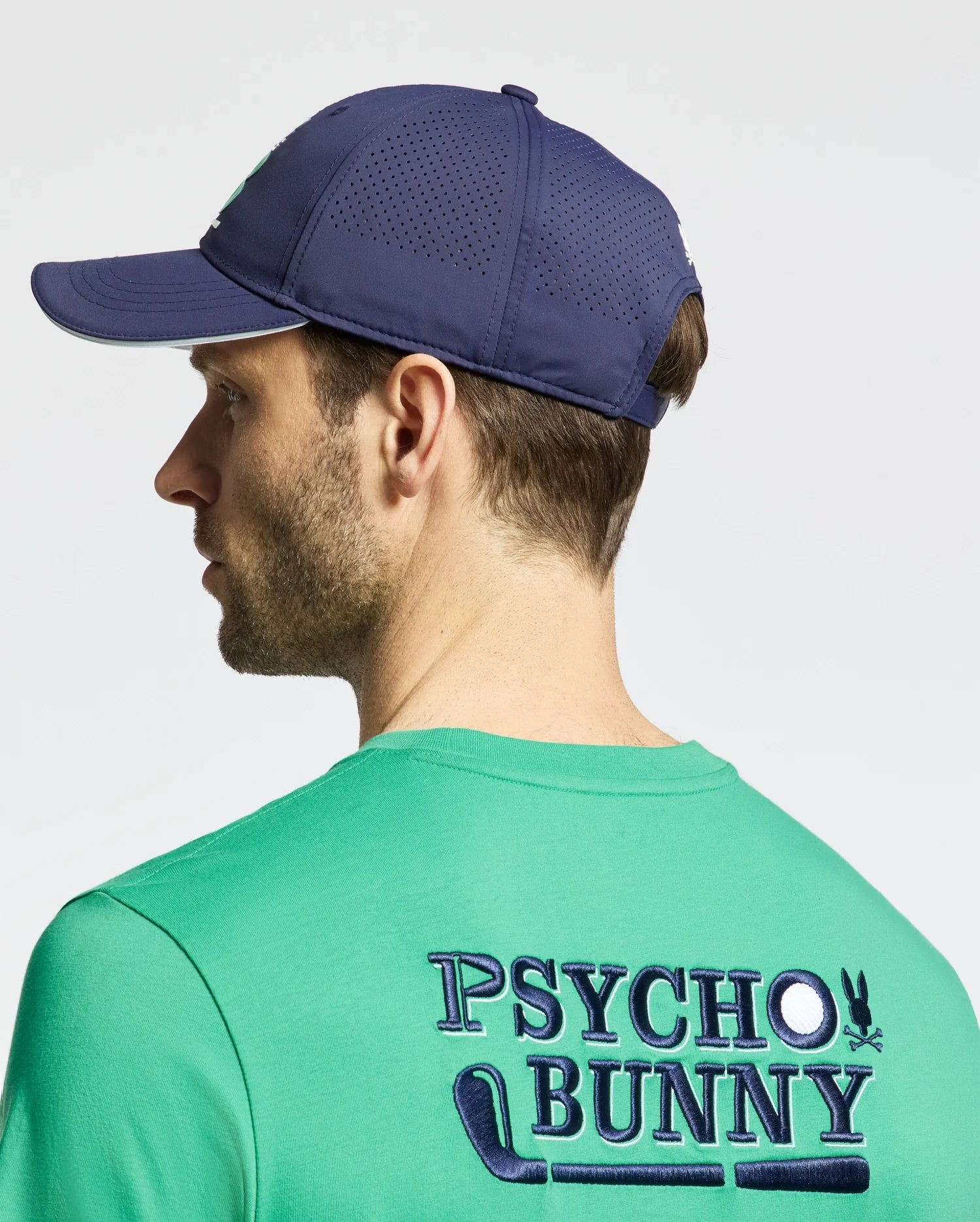 A person is shown from the back wearing a sporty green Psycho Bunny t-shirt, made of lightweight cotton blend, featuring a stylized "Psycho Bunny" logo with a bunny and golf club graphic, paired with the blue MENS DENIS SPORT CAP - B6A642F200 from Psycho Bunny.