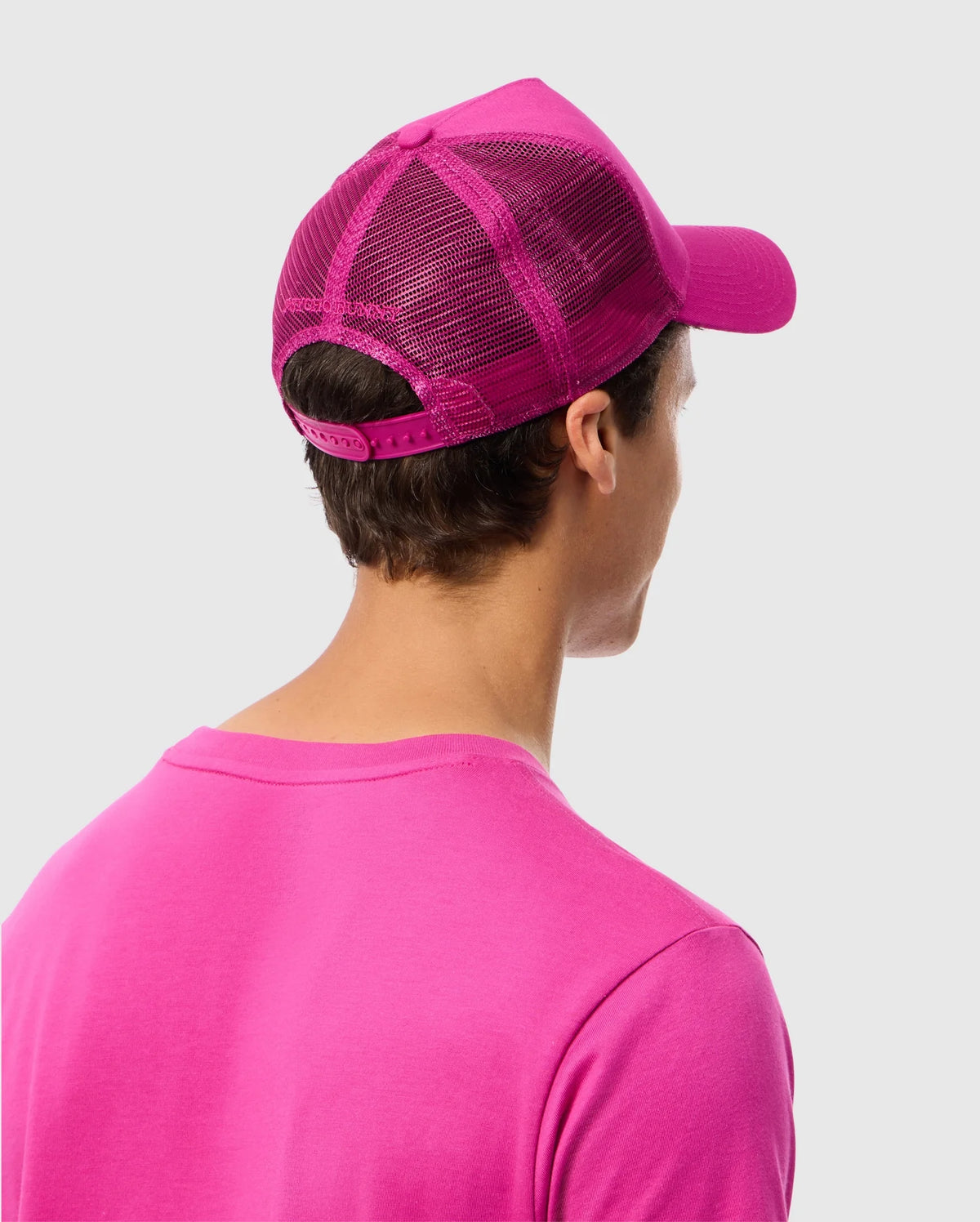 A person wearing a bright pink Psycho Bunny MENS RIVIERA TRUCKER CAP - B6A667C200 with snapback fastening and a matching pink shirt is shown from the back. The background is plain and light gray. The person's face is not visible, evoking early 2000s nostalgia.