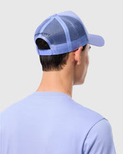 A person is seen from behind wearing a light blue Psycho Bunny MENS RIVIERA TRUCKER CAP - B6A667C200 with a mesh back and snapback fastening, along with a matching light blue t-shirt. The cap evokes early 2000s nostalgia against the plain, light-colored background.