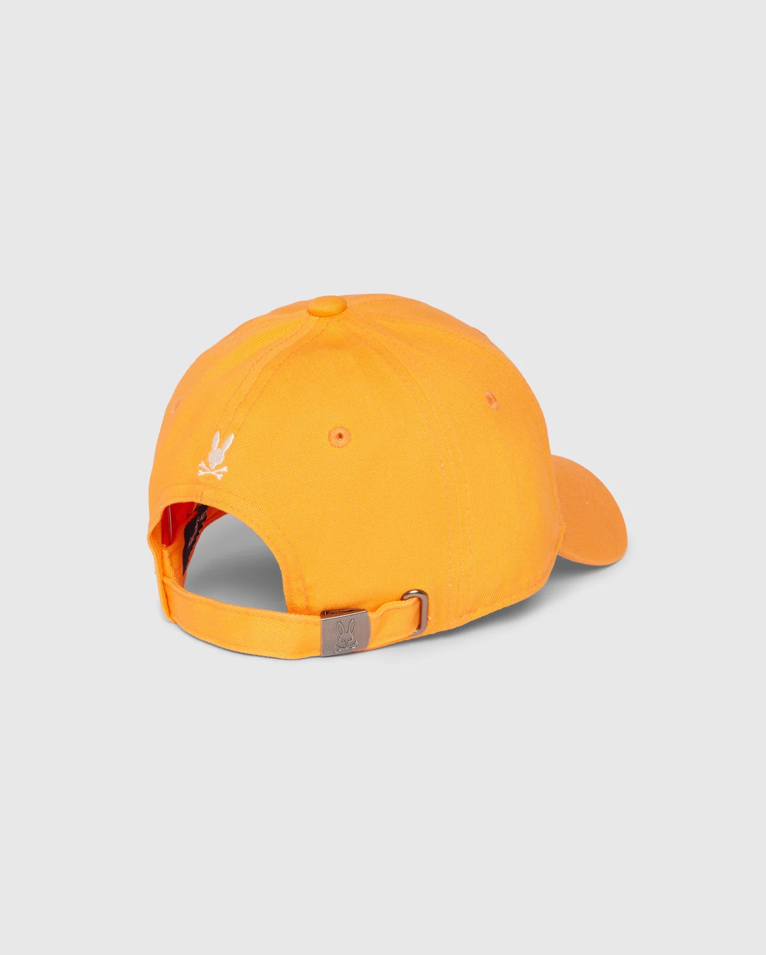Back view of the MENS KAYDEN BASEBALL CAP - B6A677C200 by Psycho Bunny, made from orange cotton twill with an adjustable strap and metal buckle. The cap showcases a small white embroidered Bunny logo above the strap and includes eyelets for ventilation. The background is plain gray.
