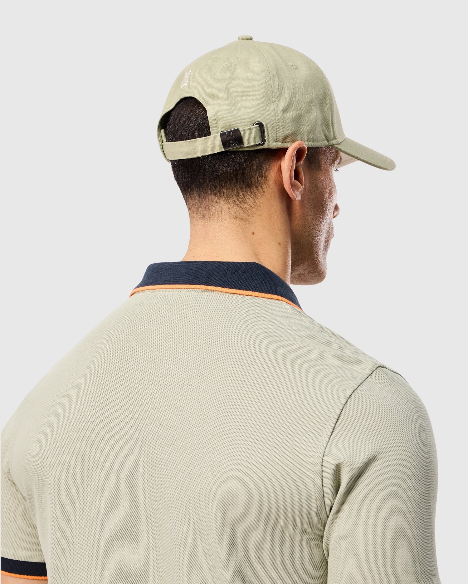Man photographed from behind wearing a light green polo shirt with dark collar and sleeve trim and a matching light green MENS KAYDEN BASEBALL CAP - B6A677C200 by Psycho Bunny. The light-colored background highlights the neat cotton twill texture of his shirt, subtly adorned with a bunny logo.