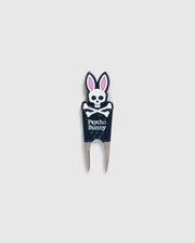 Image of a DIVOT TOOL - B6A699C200 from Psycho Bunny with a design featuring a skull with bunny ears and the text "Psycho Bunny" underneath. The tool has two prongs at the bottom, and the skull and ears are in black, white, and pink. A perfect addition to golf gifts, this tool also comes with a magnetic ball marker. The background is plain gray.