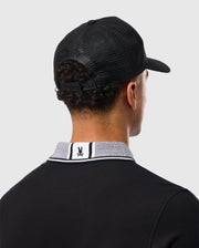 A man with short curly hair is seen from behind, wearing the MENS MALTON TRUCKER CAP - B6A797D200 by Psycho Bunny, a black trucker cap featuring mesh ventilation, paired with a 100% cotton polo shirt. The polo shirt has a white collar adorned with black and white stripes and features a small rabbit logo on the back. The background is plain light grey.