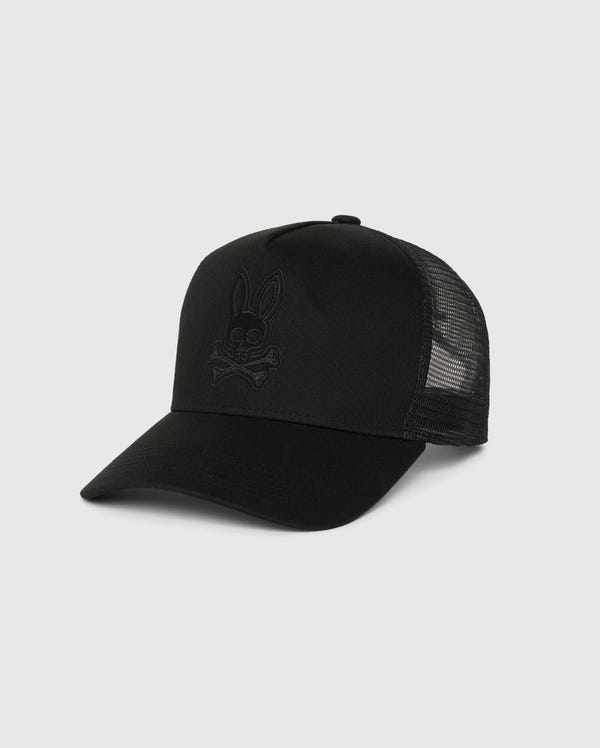The MENS MALTON TRUCKER CAP - B6A797D200 by Psycho Bunny is a black trucker cap featuring a curved brim and mesh ventilation at the back. The front panel, made of 100% cotton, showcases a dark, embroidered bunny skull and crossbones design on a plain, light gray background.