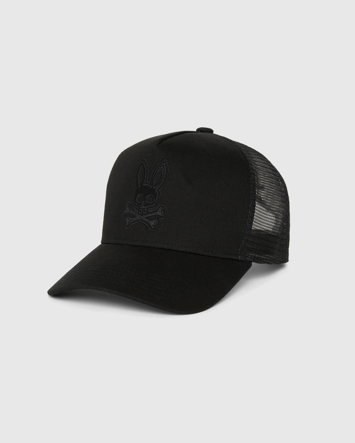 The MENS MALTON TRUCKER CAP - B6A797D200 by Psycho Bunny is a black trucker cap featuring a curved brim and mesh ventilation at the back. The front panel, made of 100% cotton, showcases a dark, embroidered bunny skull and crossbones design on a plain, light gray background.