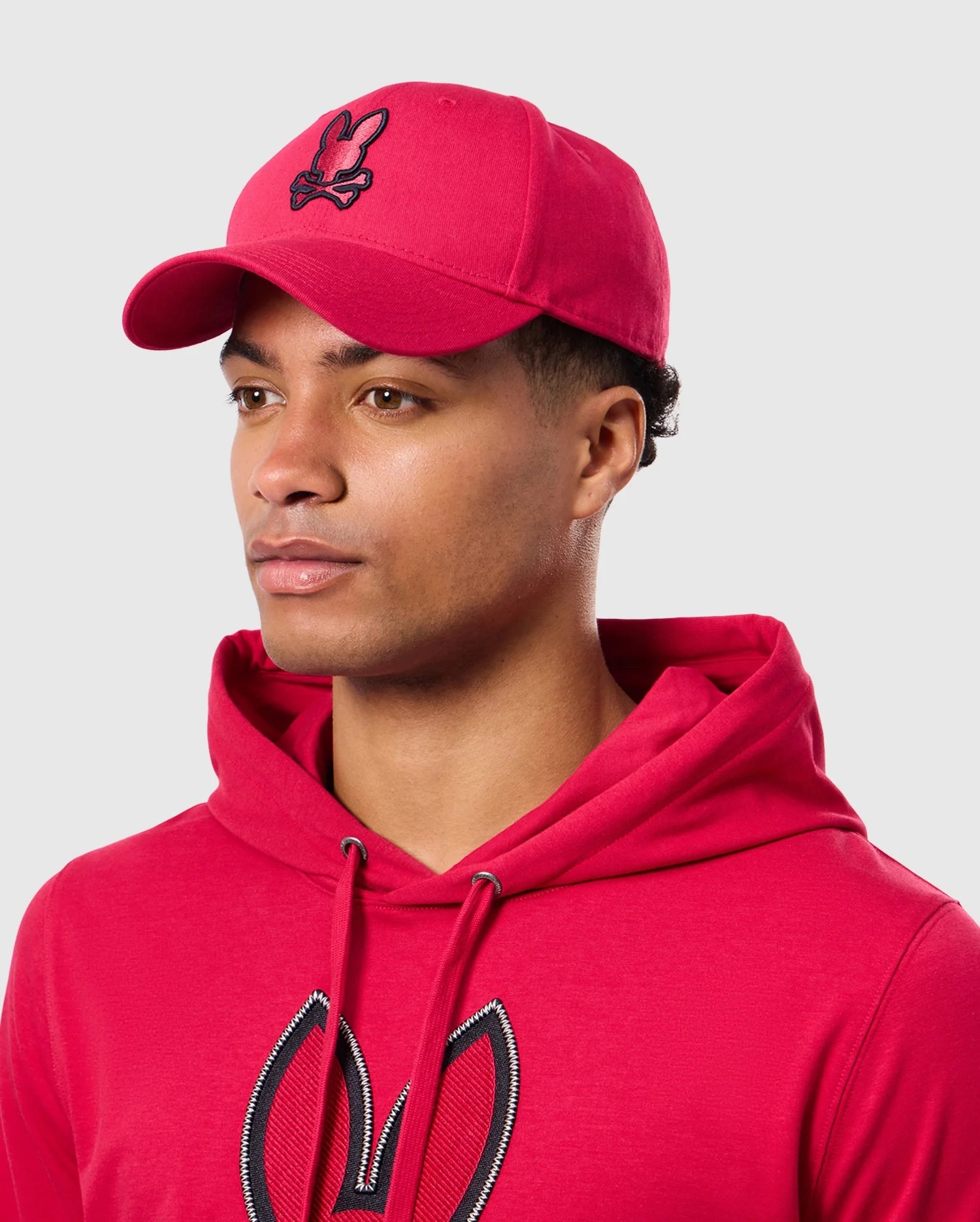 A person wearing the Psycho Bunny MENS WALTER BASEBALL CAP - B6A800D200 and a matching red hoodie with a bunny logo, looking to the side against a plain background. The durable and comfortable cap is crafted from 100% cotton, perfectly complementing the casual outfit.