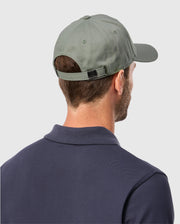 A person is shown from the back wearing a Psycho Bunny Men's Classic Baseball Cap (B6A816B200) in green and a dark polo shirt. The cap's strap highlights its solid pattern, set against a light gray background.