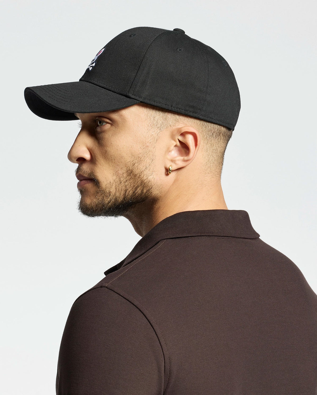 A person with a trimmed beard and short hair dons the Psycho Bunny MENS CLASSIC BASEBALL CAP - B6A816A1HT. The adjustable black cap features an embroidered Bunny logo and pairs well with a dark polo shirt as the individual faces left against a plain, light-colored background.