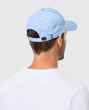 A person is wearing a Psycho Bunny MENS CLASSIC BASEBALL CAP - B6A816B200 in light blue and a white t-shirt, facing away from the camera. The 100% cotton cap includes an adjustable strap at the back. The background is simple and light gray.