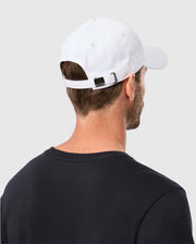 A person is seen from behind wearing a classic black t-shirt and a Psycho Bunny MENS CLASSIC BASEBALL CAP - B6A816A1HT in white cotton, which includes an adjustable strap at the back. The individual stands against a plain, light gray background.