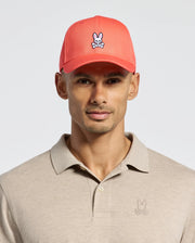 A person wears a beige Polo shirt with a bunny logo and Psycho Bunny's red MENS CLASSIC BASEBALL CAP - B6A816F200, crafted from cotton twill, featuring the bunny logo. The background is plain white.