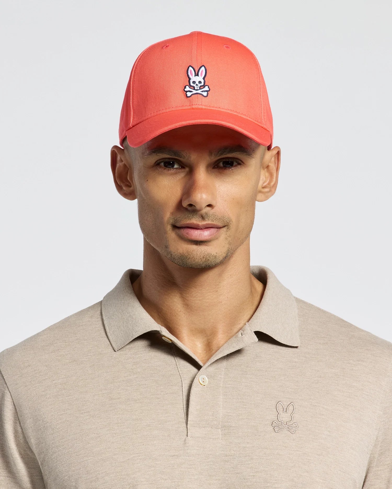 A person wears a beige Polo shirt with a bunny logo and Psycho Bunny's red MENS CLASSIC BASEBALL CAP - B6A816F200, crafted from cotton twill, featuring the bunny logo. The background is plain white.