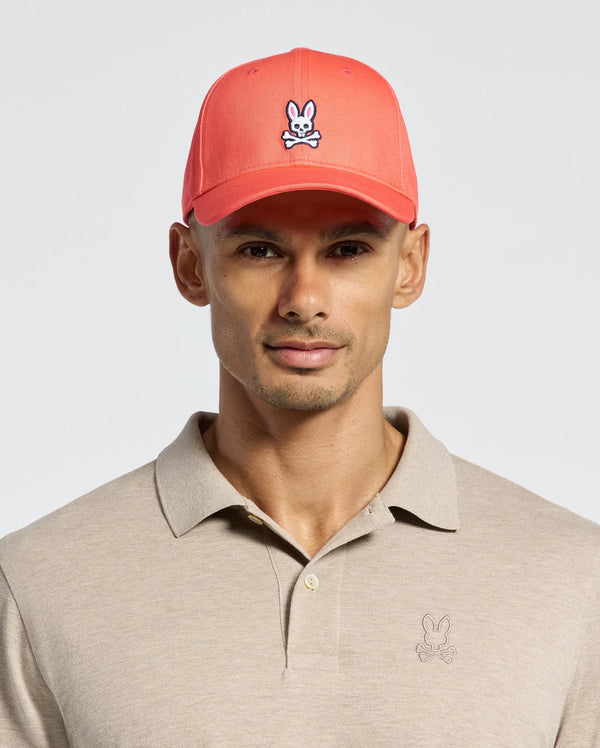 A person wears a beige Polo shirt with a bunny logo and Psycho Bunny's red MENS CLASSIC BASEBALL CAP - B6A816F200, crafted from cotton twill, featuring the bunny logo. The background is plain white.