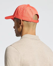 A person is shown from the side and back wearing a beige shirt and Psycho Bunny's MENS CLASSIC BASEBALL CAP in bright coral (B6A816F200), featuring a visible adjustable fit strap. The background is plain and light-colored.