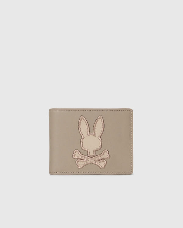 The Psycho Bunny MENS LEATHER WALLET - B6A868D200, featured on a plain background, displays an embossed rabbit's head with crossed bones. This beige wallet offers a minimalist and stylish design for those who value refined craftsmanship.