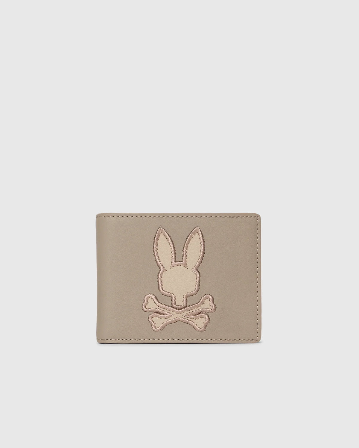 The Psycho Bunny MENS LEATHER WALLET - B6A868D200, featured on a plain background, displays an embossed rabbit's head with crossed bones. This beige wallet offers a minimalist and stylish design for those who value refined craftsmanship.