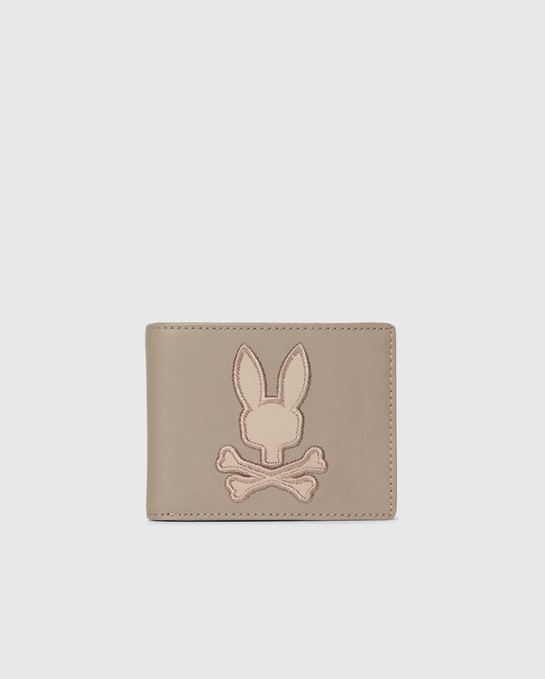 The Psycho Bunny MENS LEATHER WALLET - B6A868D200, featured on a plain background, displays an embossed rabbit's head with crossed bones. This beige wallet offers a minimalist and stylish design for those who value refined craftsmanship.
