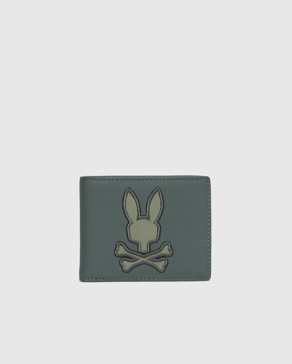 Introducing the MENS WORDMARK LEATHER WALLET - B6A868D200 by Psycho Bunny, a stylish accessory for men. This trendy green leather wallet showcases an embroidered rabbit head above crossbones on the front, set against a sleek white background.