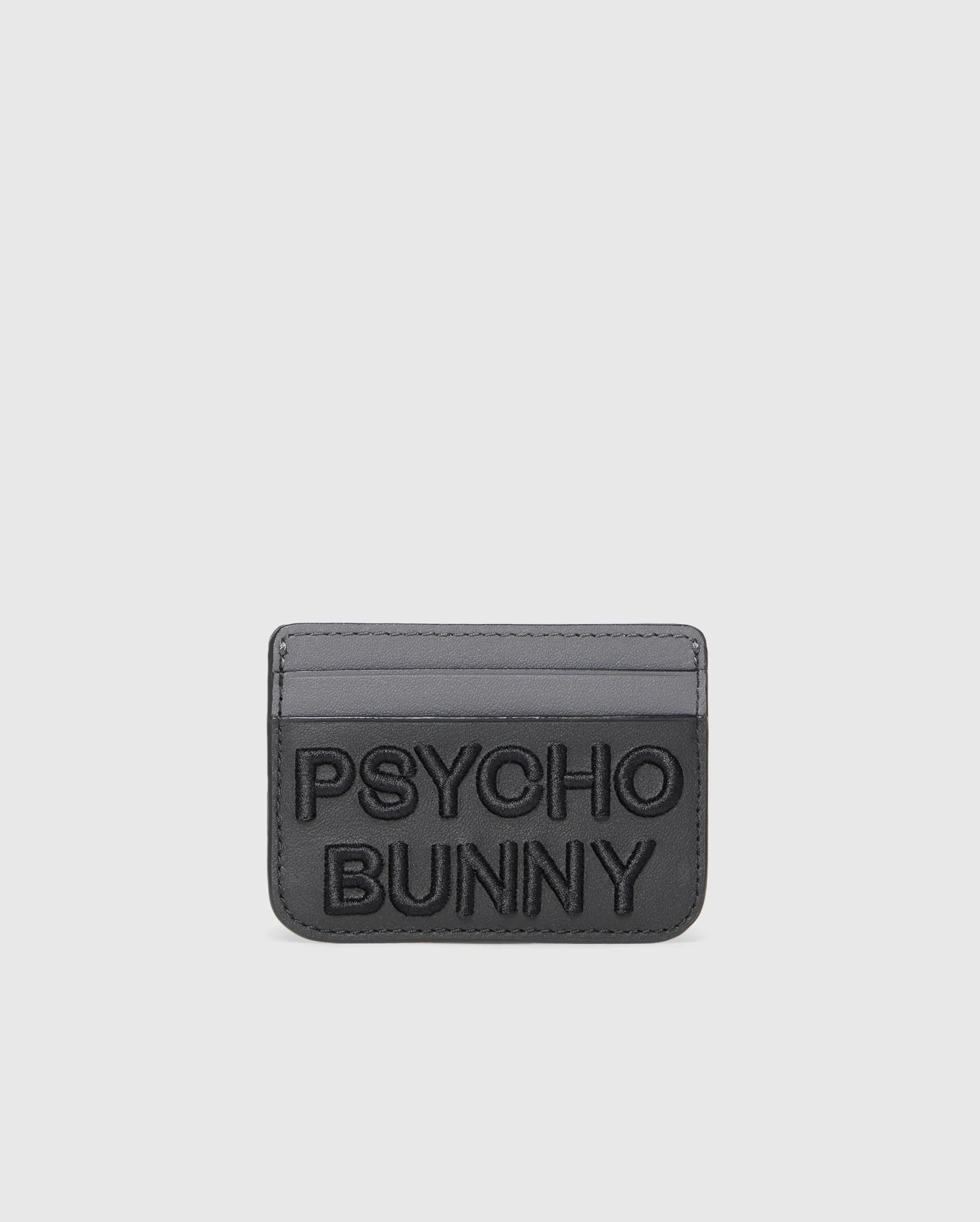 The MENS LEATHER CARD CASE - B6A869D200 by Psycho Bunny is a gray leather card holder with embossed "PSYCHO BUNNY" text on the front and two card slots against a sleek white backdrop. Crafted from premium leather, it offers a minimalist design.