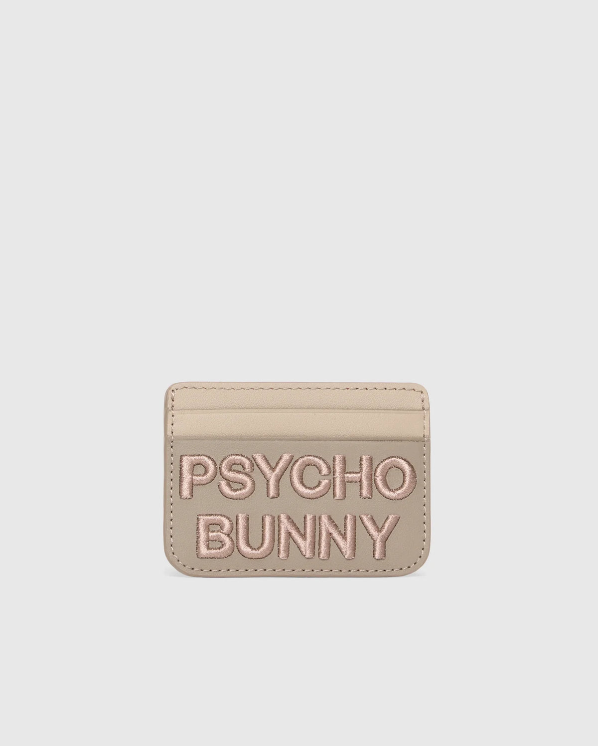 A premium beige leather cardholder features "PSYCHO BUNNY" embroidered in pink on the front. The minimalist MENS LEATHER CARD CASE - B6A869D200 by Psycho Bunny offers style and simplicity, set against a plain white background.