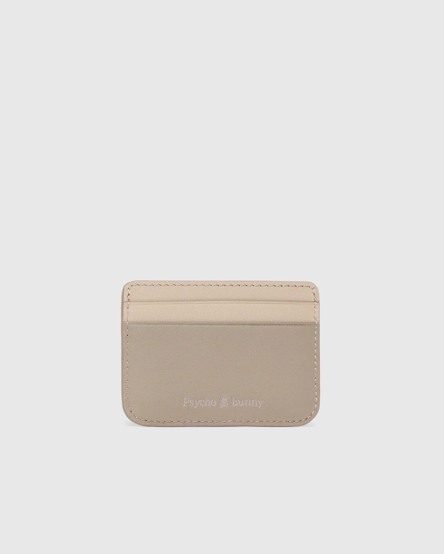 The MENS LEATHER CARD CASE - B6A869D200 by Psycho Bunny is a compact light beige leather card holder with two slots and an elegant "Psycho Bunny" embossing on the lower front. Photographed against a white background, it highlights the sleek design.