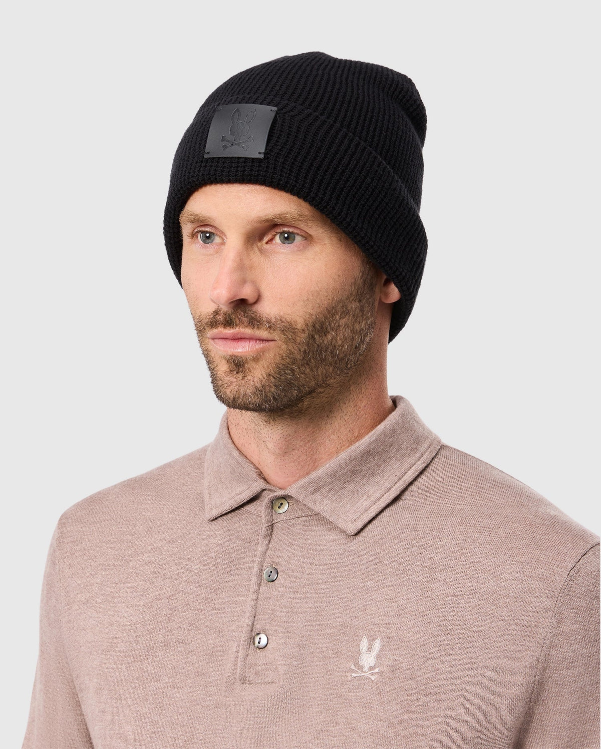 A man wearing the Psycho Bunny MENS WAFFLE BEANIE - B6A873D200 in black and a light brown cotton knit sweater featuring a small embroidered logo. He has a beard and is gazing slightly to the side against a plain background.