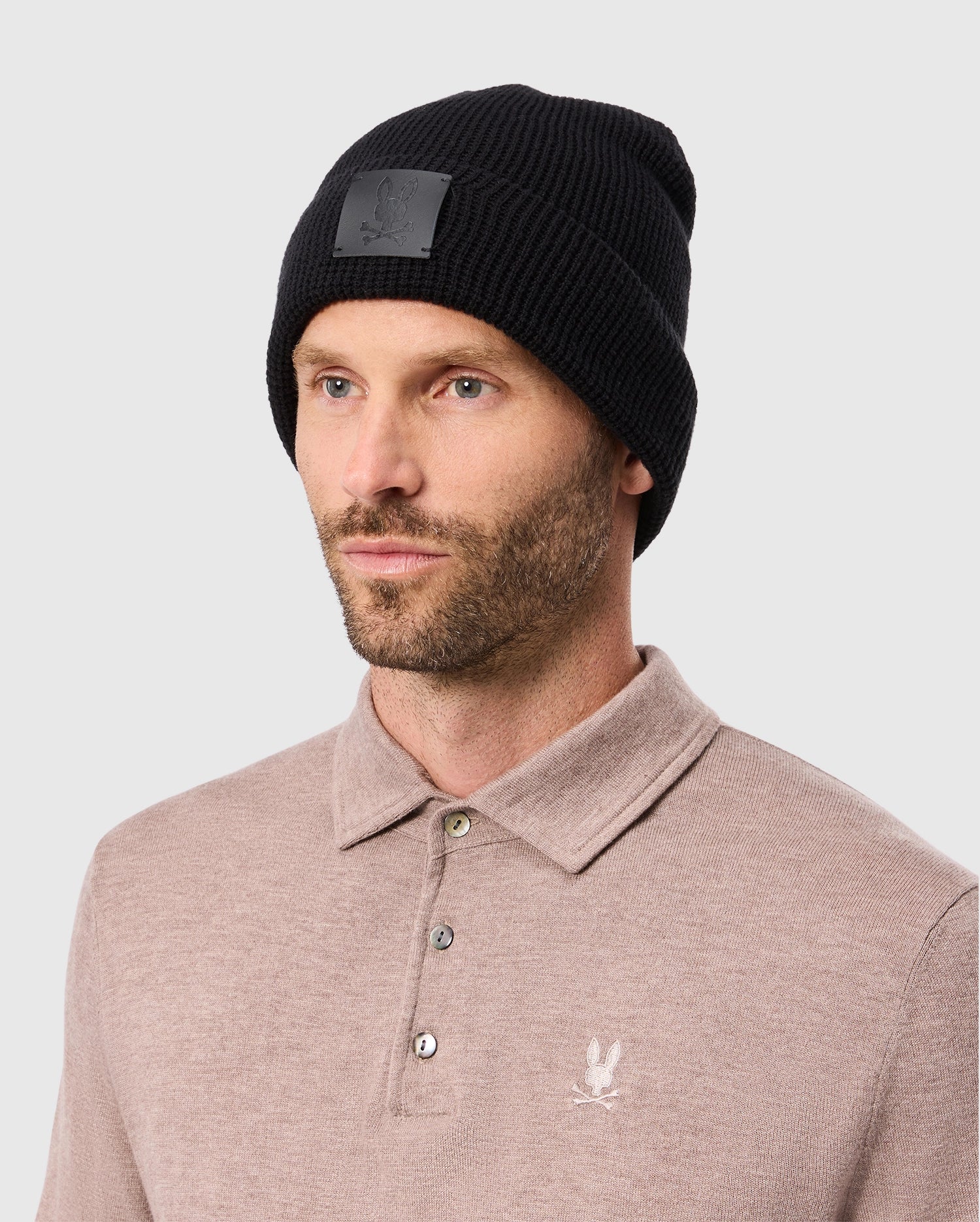 A man wearing the Psycho Bunny MENS WAFFLE BEANIE - B6A873D200 in black and a light brown cotton knit sweater featuring a small embroidered logo. He has a beard and is gazing slightly to the side against a plain background.