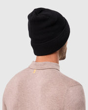 Rear view of a person wearing a cozy beige sweater and the Psycho Bunny MENS WAFFLE BEANIE - B6A873D200 in black, standing against a plain light gray background.