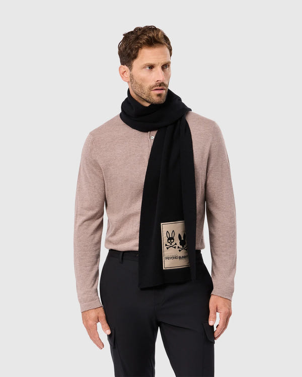 A man wearing a beige long-sleeve shirt stands against a gray background, elegantly adorned with Psycho Bunny's luxury MENS MONROE TWILL PATCH SCARF (B6A892D200) crafted from a black cotton wool blend. The scarf is distinguished by a charming bunny design patch. He has short dark hair and a beard, looking to his left.
