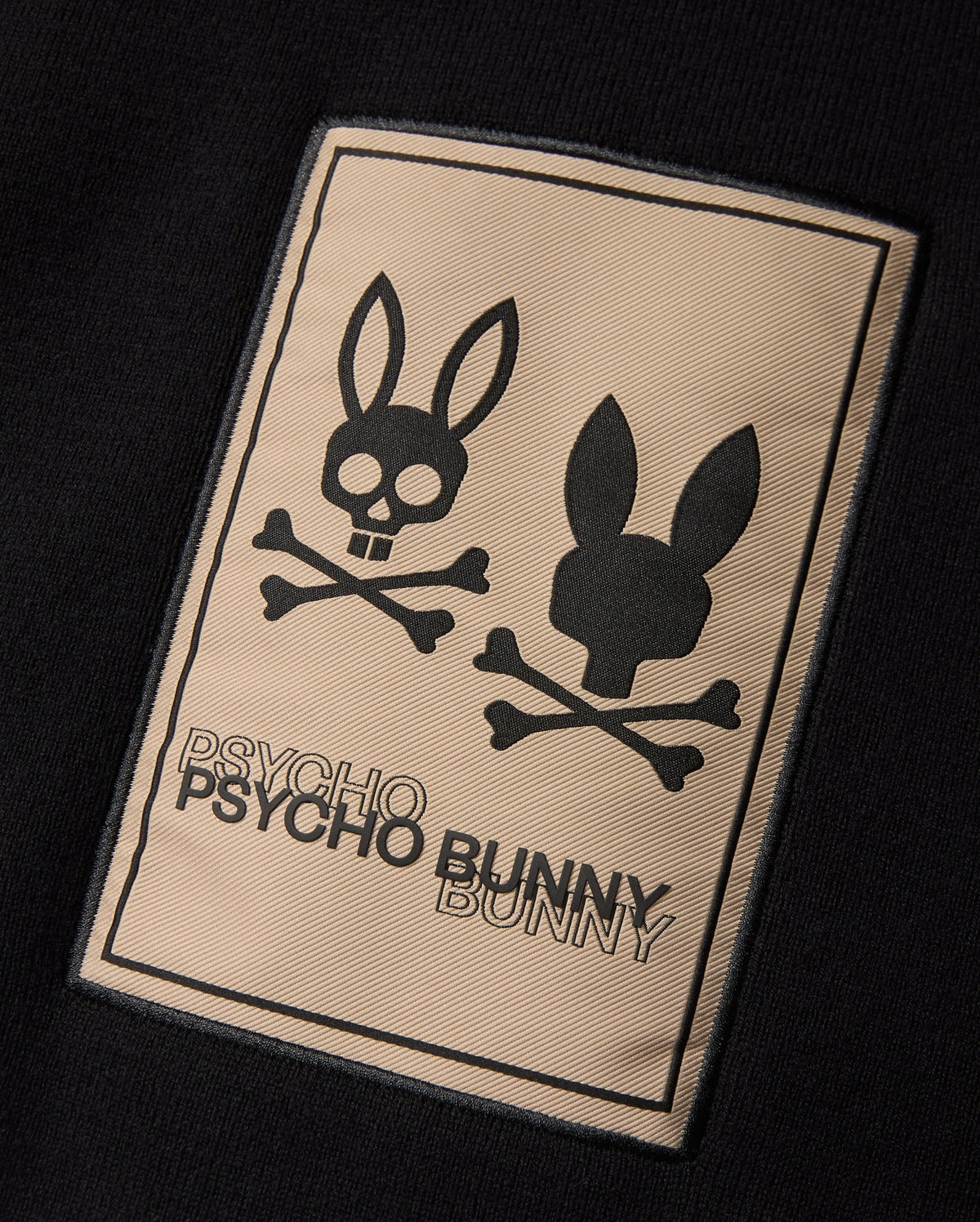 The MENS MONROE TWILL PATCH SCARF by Psycho Bunny features a luxurious black cotton wool blend. It boasts a rectangular patch embellished with two bunny head skull and crossbones designs. The brand name "Psycho Bunny" is elegantly emblazoned in bold lettering below, enhancing its edgy sophistication.