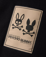 The MENS MONROE TWILL PATCH SCARF by Psycho Bunny features a luxurious black cotton wool blend. It boasts a rectangular patch embellished with two bunny head skull and crossbones designs. The brand name "Psycho Bunny" is elegantly emblazoned in bold lettering below, enhancing its edgy sophistication.