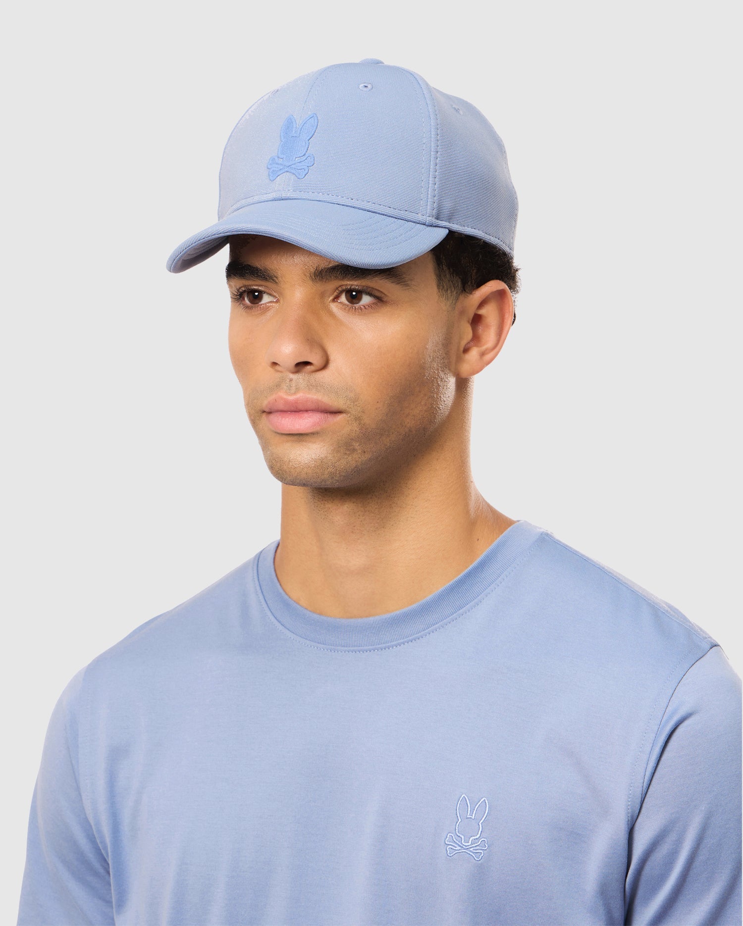 A person stands against a white background wearing a light blue Psycho Bunny MENS JAMES PREMIUM BASEBALL CAP - B6A953D200 and a matching T-shirt with a tonal embroidered logo, crafted from knit twill fabric.