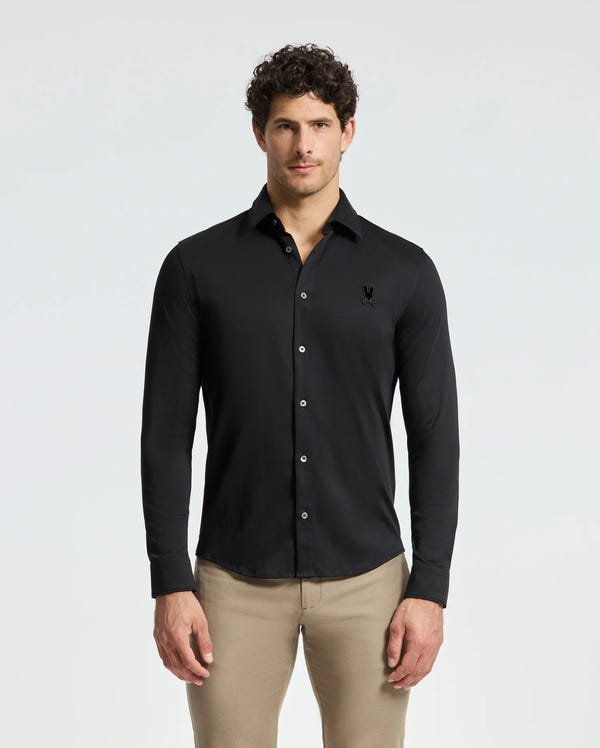 A man with curly hair wears a Psycho Bunny Men's Interlock Knit Shirt in black, crafted from 100% Pima Cotton, stylishly paired with beige pants. He stands confidently against a plain white background, projecting a classic and refined look.