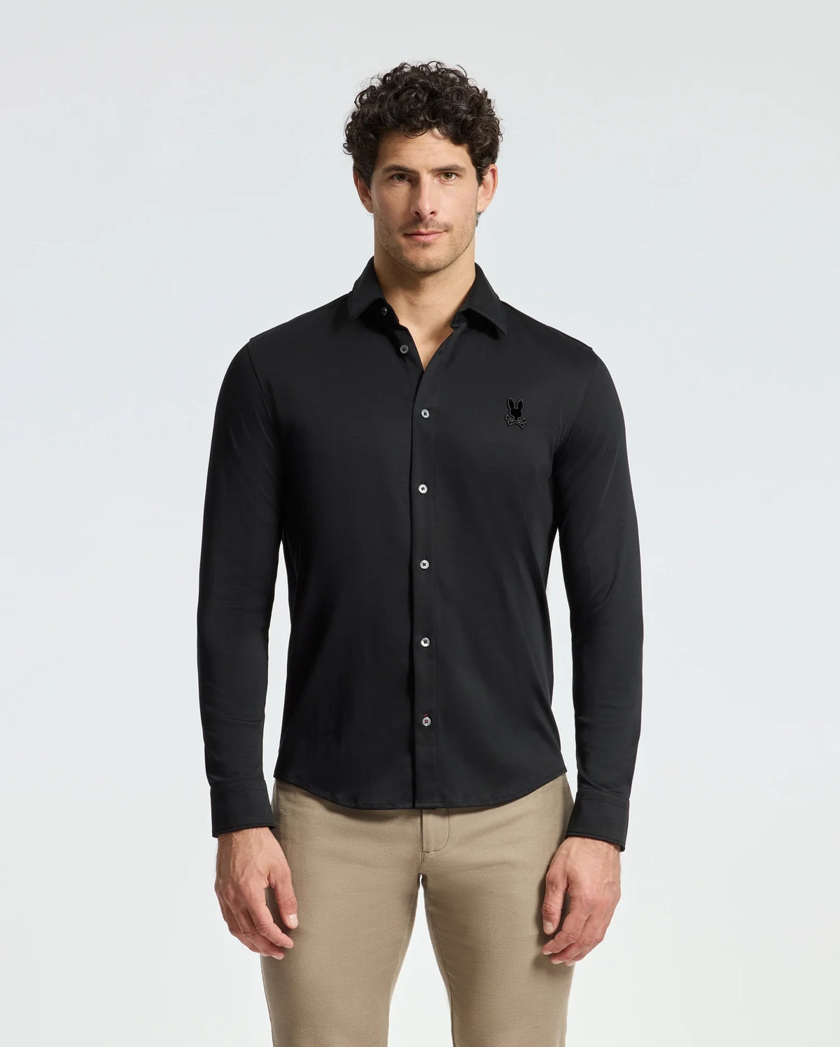 A man with curly hair wears a Psycho Bunny Men's Interlock Knit Shirt in black, crafted from 100% Pima Cotton, stylishly paired with beige pants. He stands confidently against a plain white background, projecting a classic and refined look.