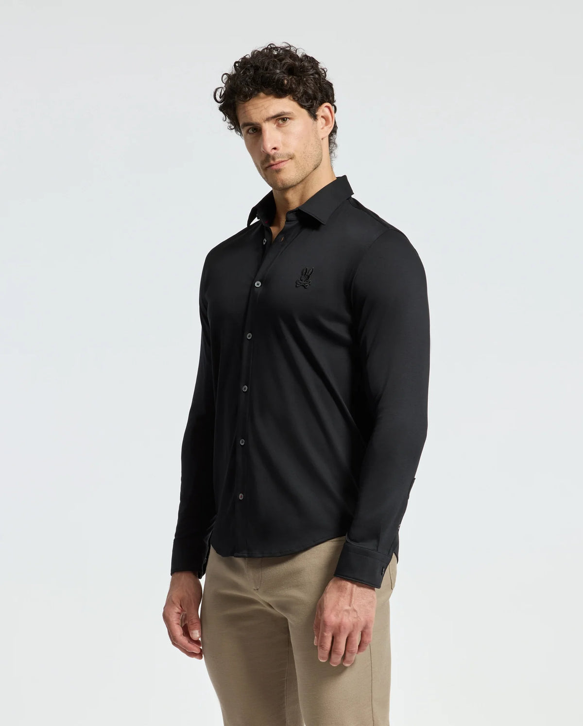 A man with curly hair wears a fitted men’s black knit shirt from Psycho Bunny, specifically the MENS INTERLOCK KNIT SHIRT - B6C188E200, made from 100% Pima cotton. He pairs it with beige pants and stands against a plain white background, looking towards the camera with a neutral expression.
