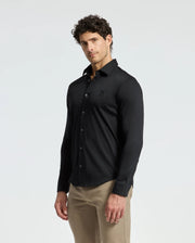 A man with curly hair wears a fitted men’s black knit shirt from Psycho Bunny, specifically the MENS INTERLOCK KNIT SHIRT - B6C188E200, made from 100% Pima cotton. He pairs it with beige pants and stands against a plain white background, looking towards the camera with a neutral expression.