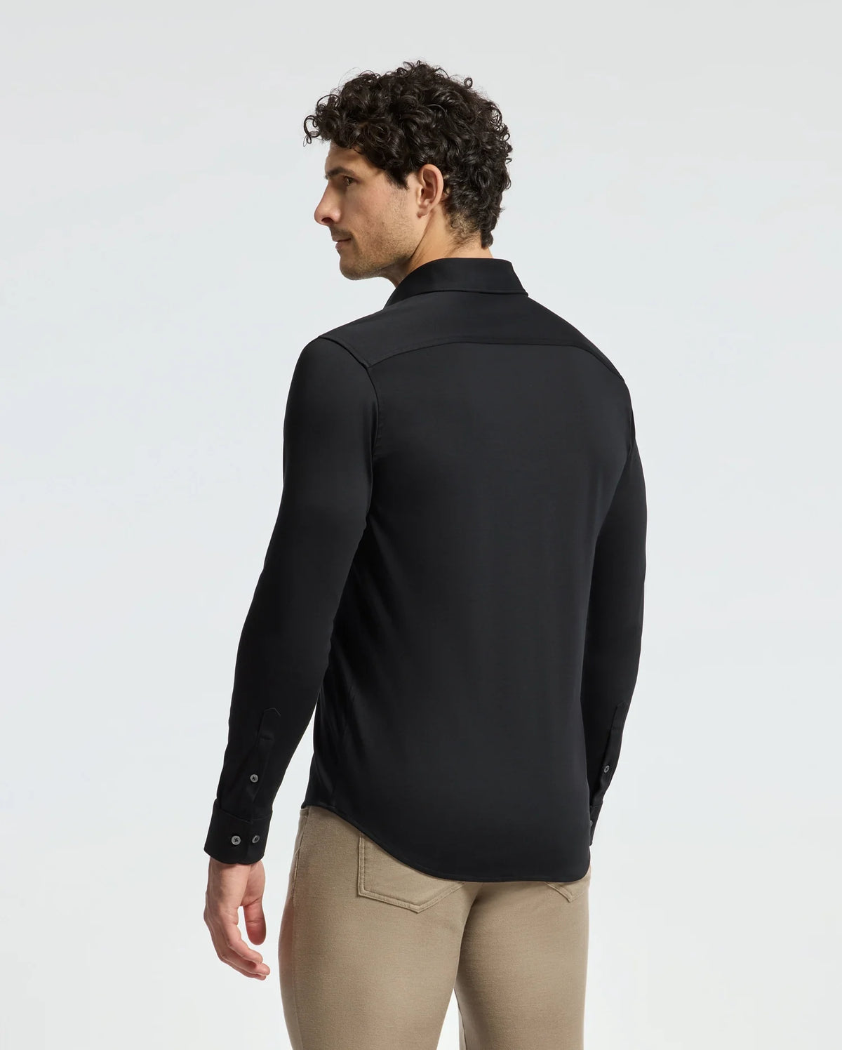 A man with curly hair is wearing the Psycho Bunny Men's Interlock Knit Shirt (B6C188E200) in black, paired with beige pants, while gazing to the left against a plain background. Made from 100% Pima cotton, his shirt offers a sophisticated and comfortable feel, ideal for any occasion.