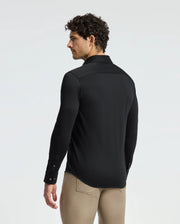 A man with curly hair is wearing the Psycho Bunny Men's Interlock Knit Shirt (B6C188E200) in black, paired with beige pants, while gazing to the left against a plain background. Made from 100% Pima cotton, his shirt offers a sophisticated and comfortable feel, ideal for any occasion.