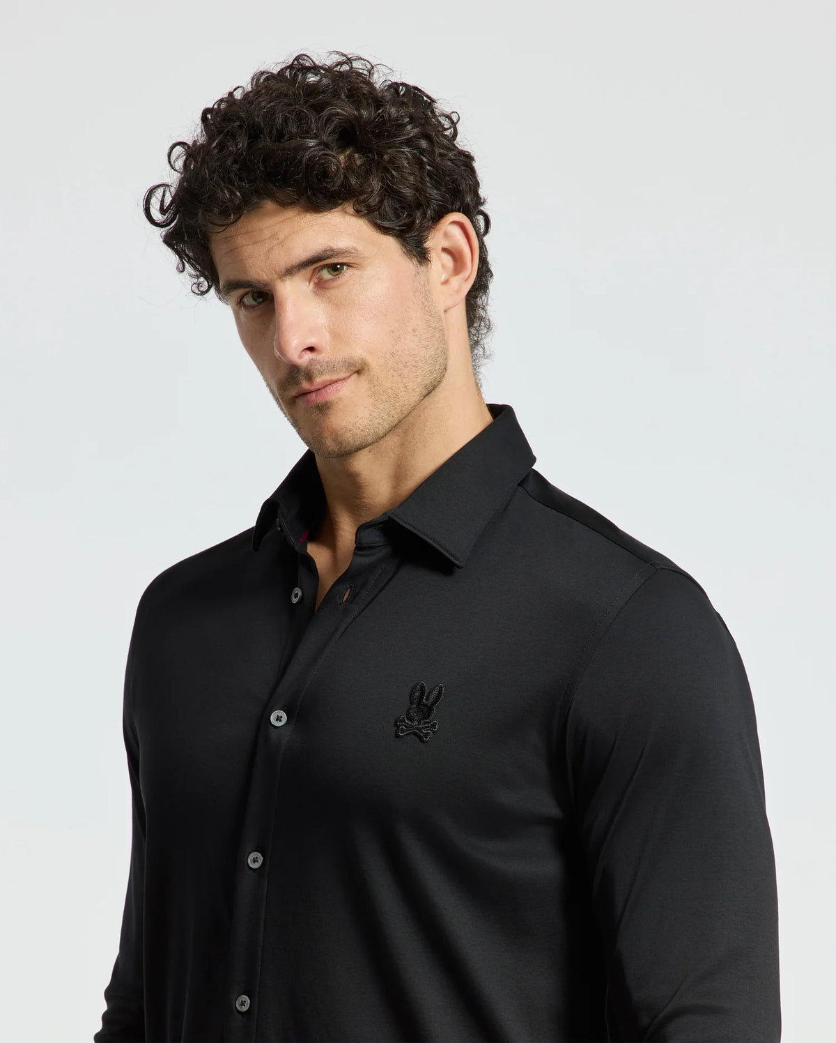 A man with curly hair is wearing a sleek Men's Interlock Knit Shirt by Psycho Bunny, crafted from 100% Pima cotton, with a small embroidered logo on the left side. He poses confidently against a plain white background.