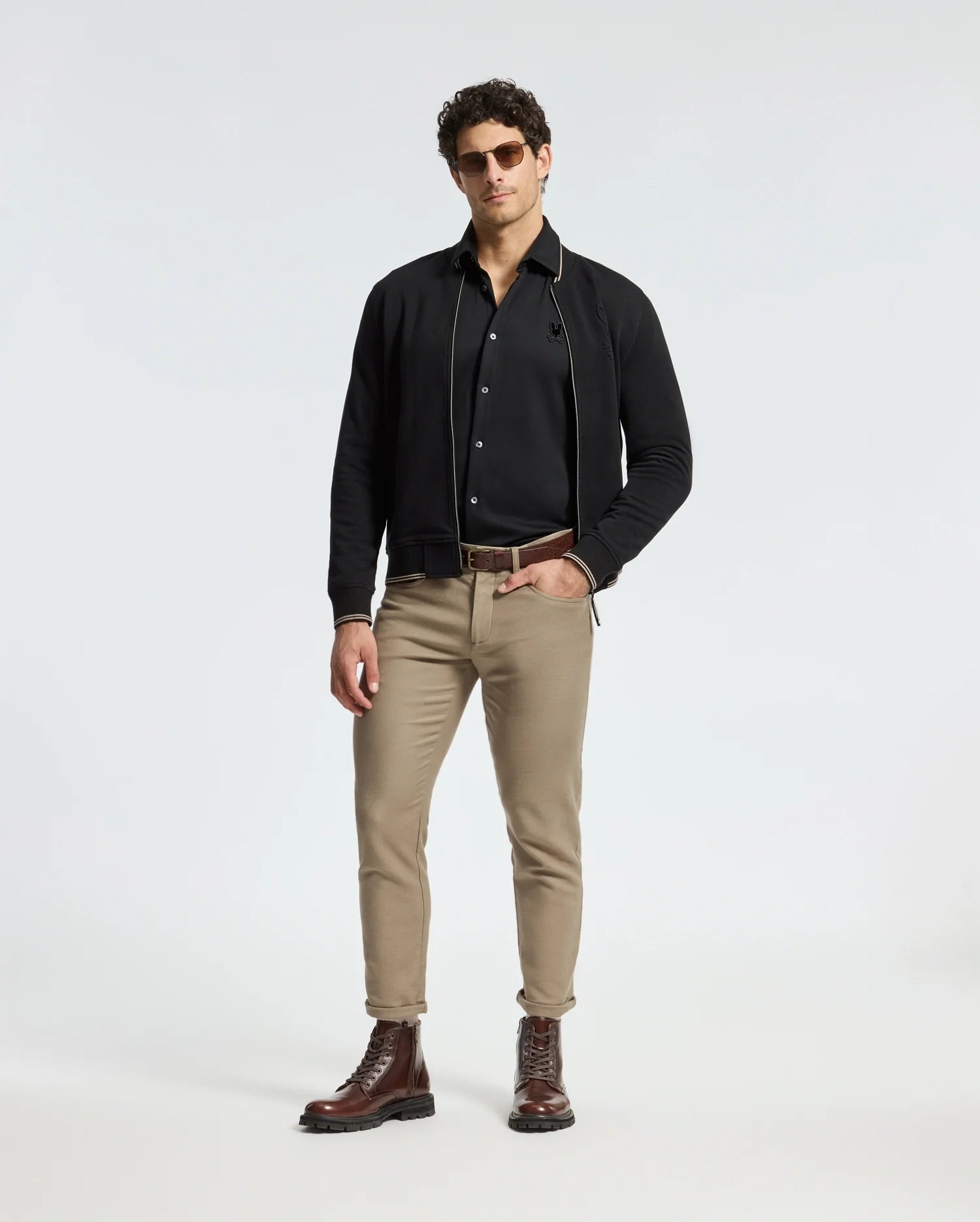 A man stands confidently wearing Psycho Bunny's MENS INTERLOCK KNIT SHIRT - B6C188E200, a sleek black collared shirt made from 100% Pima Cotton. He pairs it with a jacket and beige pants, completing the look with brown leather boots and a belt in front of a plain white background.