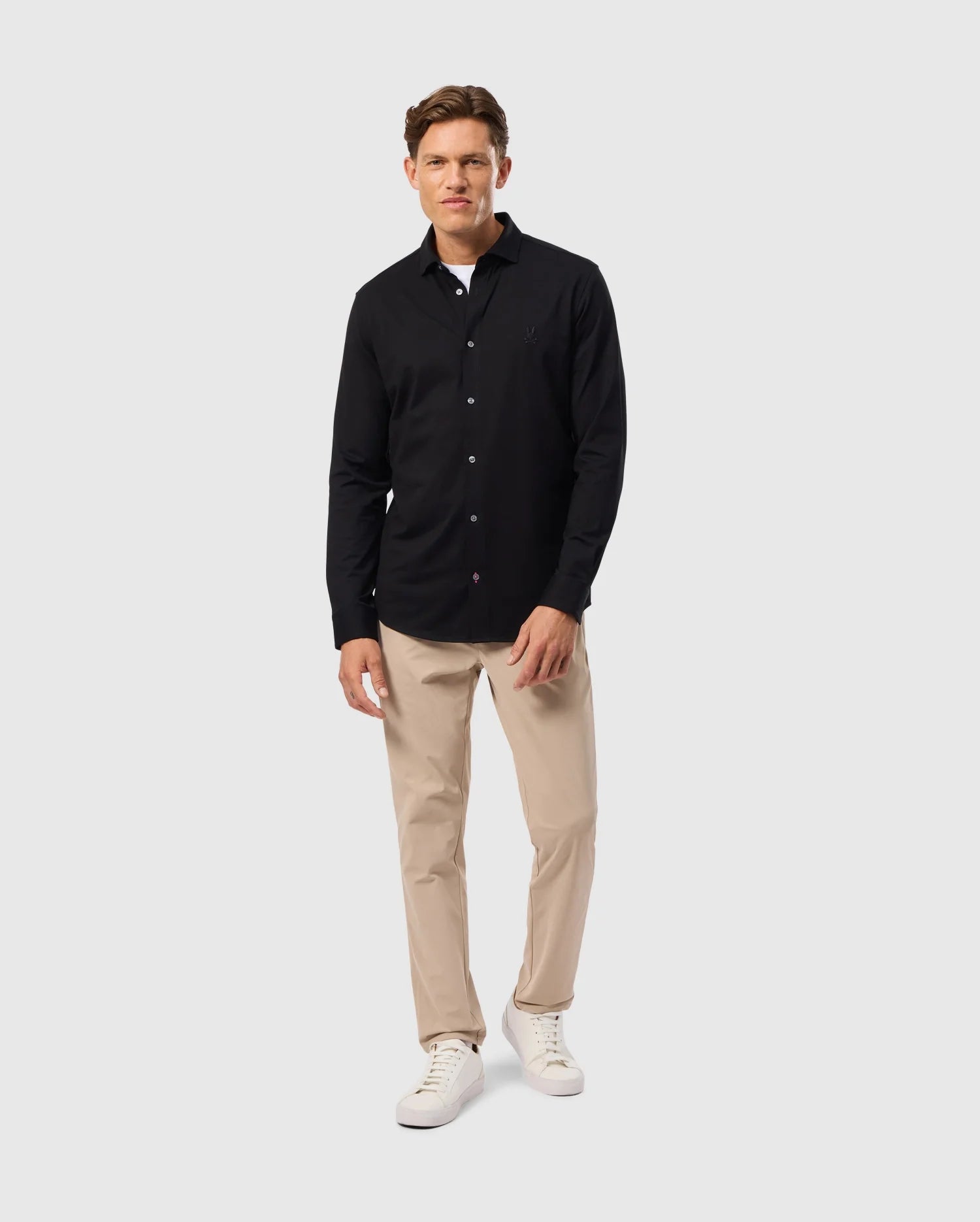 A man in a Psycho Bunny MENS ARTHUR JERSEY SHIRT stands against a light gray background. Wearing beige pants and white sneakers, he exudes a relaxed vibe with one hand in his pocket.