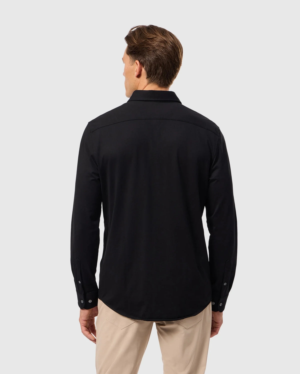 A man with short brown hair is turned away from the camera, highlighting a relaxed style in the Psycho Bunny MENS ARTHUR JERSEY SHIRT - B6C748D200, a long-sleeved black button-up crafted from Pima cotton, paired with beige pants, all set against a plain light gray backdrop.