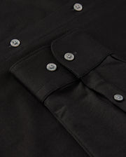 Close-up of a black MENS ARTHUR JERSEY SHIRT by Psycho Bunny, meticulously crafted from comfortable shirting and featuring a buttoned flap pocket. The shirt is neatly arranged, showcasing the fabric's texture and shiny black buttons.