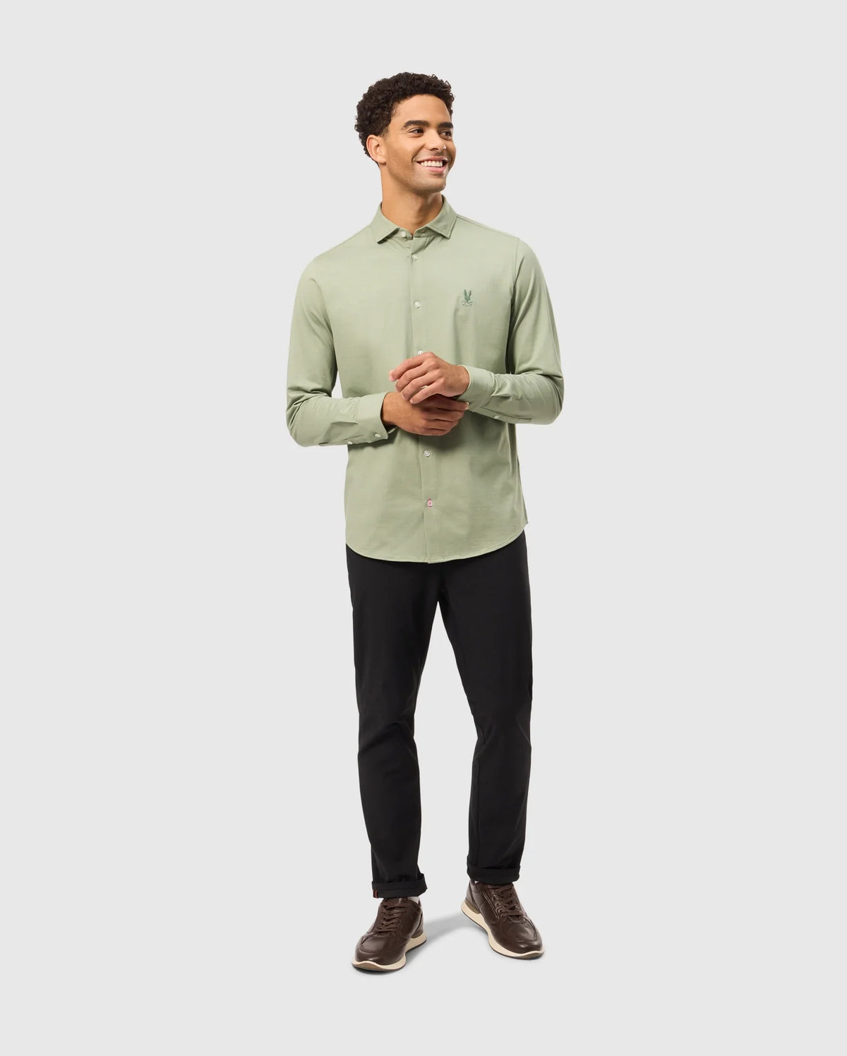 A man wearing a light green Psycho Bunny MENS BRENDAN JERSEY SHIRT, featuring an embroidered chest Bunny logo, paired with black pants, stands smiling with his hands clasped. He is also wearing brown shoes made from Pima cotton jersey. The background is plain and light gray.