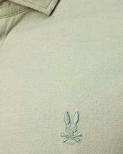 A close-up of the textured Pima cotton jersey from the Psycho Bunny MENS BRENDAN JERSEY SHIRT - B6C749D200 in light green highlights the embroidered chest Bunny logo, featuring a rabbit head with crossed bones beneath it. The design is elevated by subtle contrast piping that adds an extra touch of style.