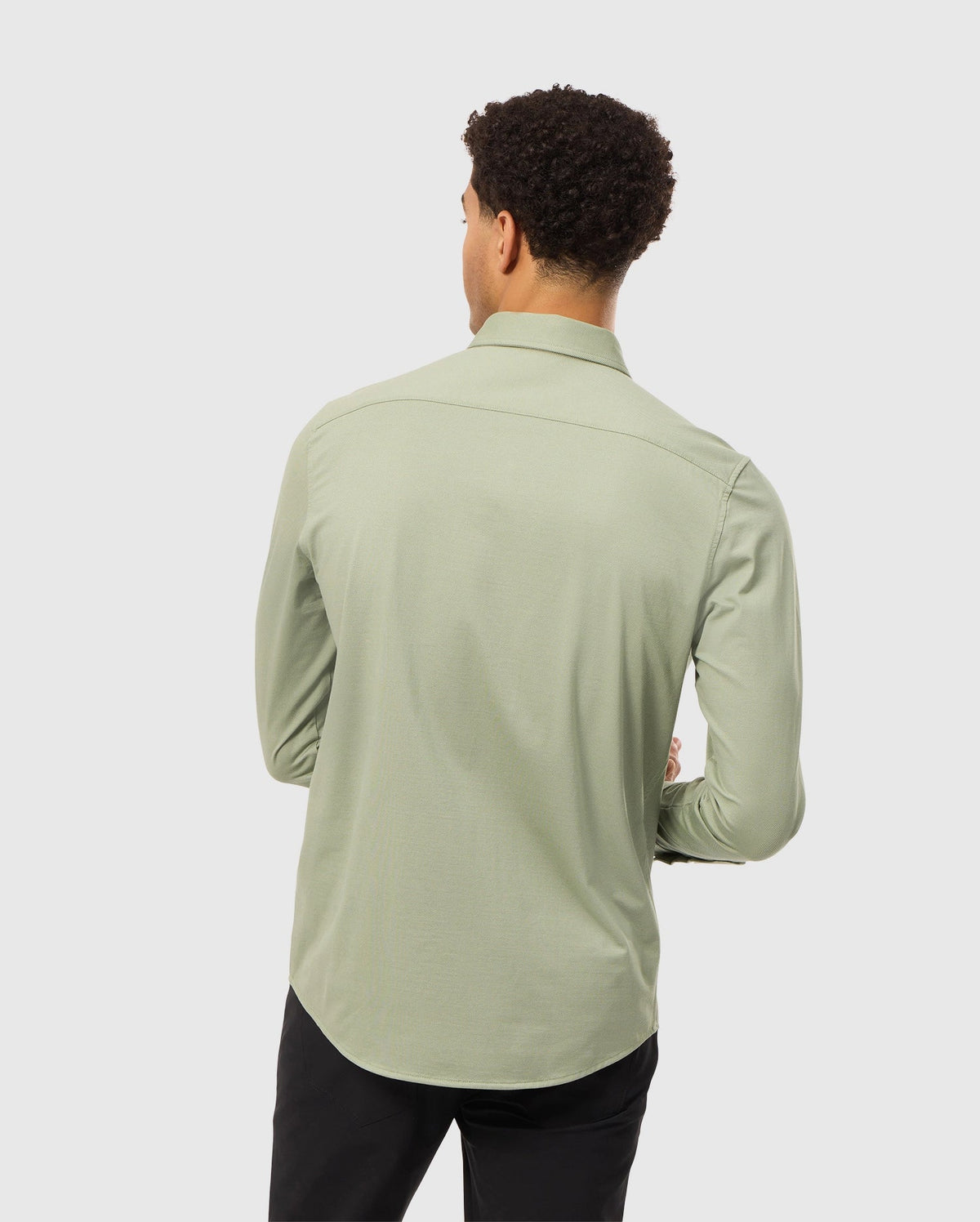 A person with short curly hair is wearing the light green MENS BRENDAN JERSEY SHIRT by Psycho Bunny, which is crafted from soft Pima cotton jersey. They are also wearing black pants and are facing away from the camera against a plain background.
